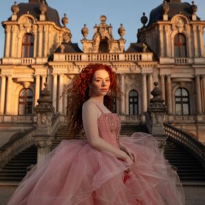 Fotoshooting, authentische Frau mit langen roten Locken in einem riesigen rosa Kleid, die in die Kamera blickt und vor einem Barockschloss steht, goldene Abendstunde